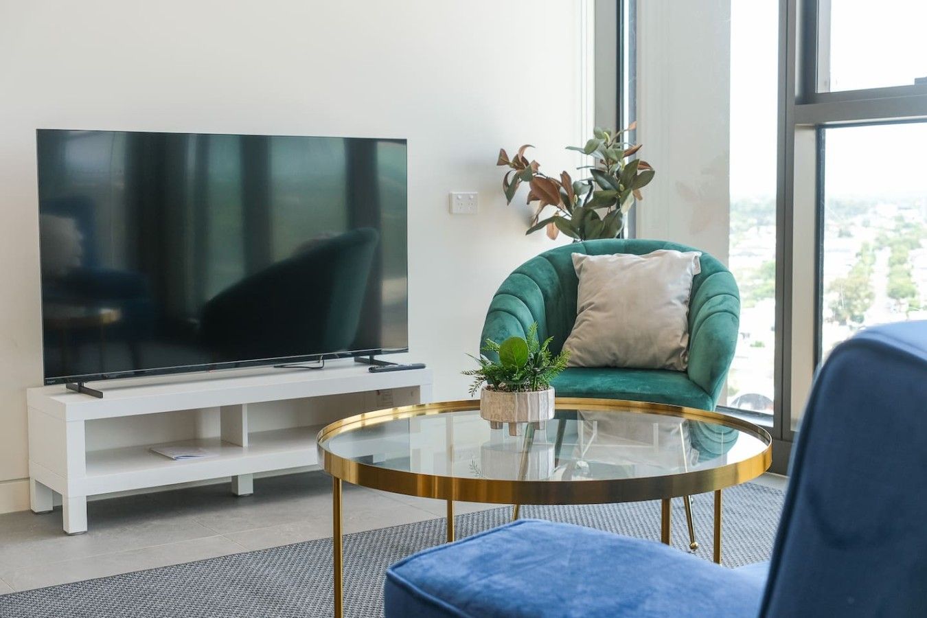 A modern living room with a flat-screen TV, a green sofa, a round glass coffee table, and a potted plant.