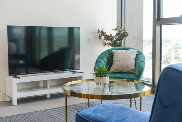 A modern living room with a flat-screen TV, a green sofa, a round glass coffee table, and a potted plant.