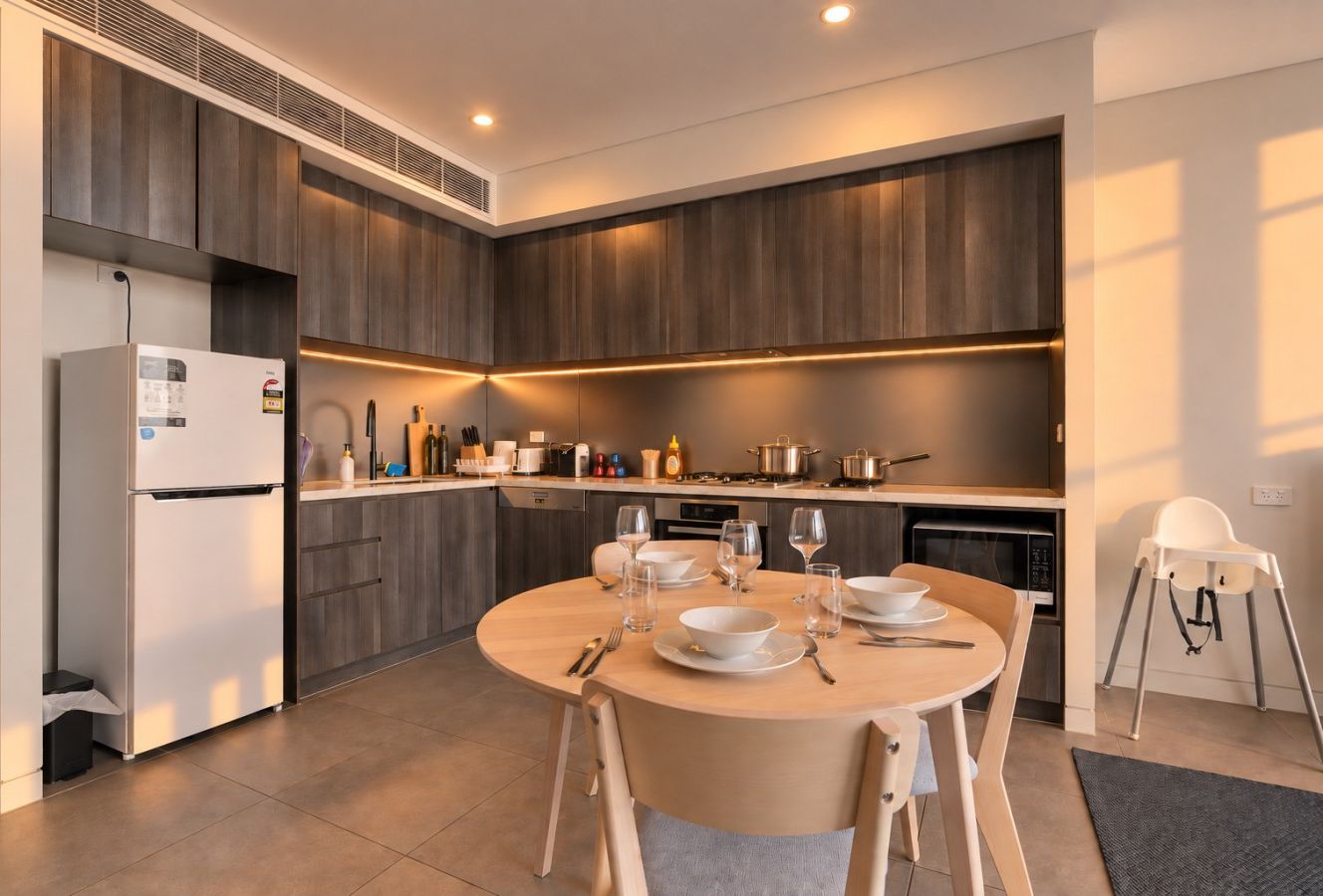 The kitchen from the dining area, showing a full size white fridge, dark cabinetry, a four burner gas cooktop, a built in oven, and a countertop microwave. The round dining table is set for four, and a white high chair is at right.