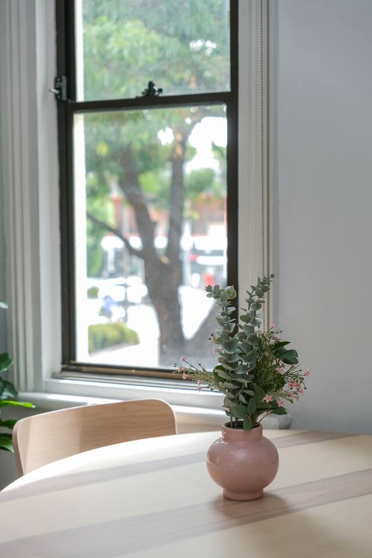 Une table avec un vase de fleurs et une fenêtre donnant sur des arbres et des bâtiments.