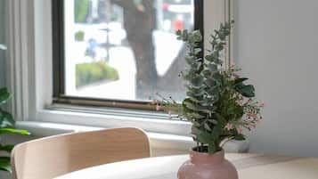A table with a vase of flowers and a window with a view of trees and buildings.