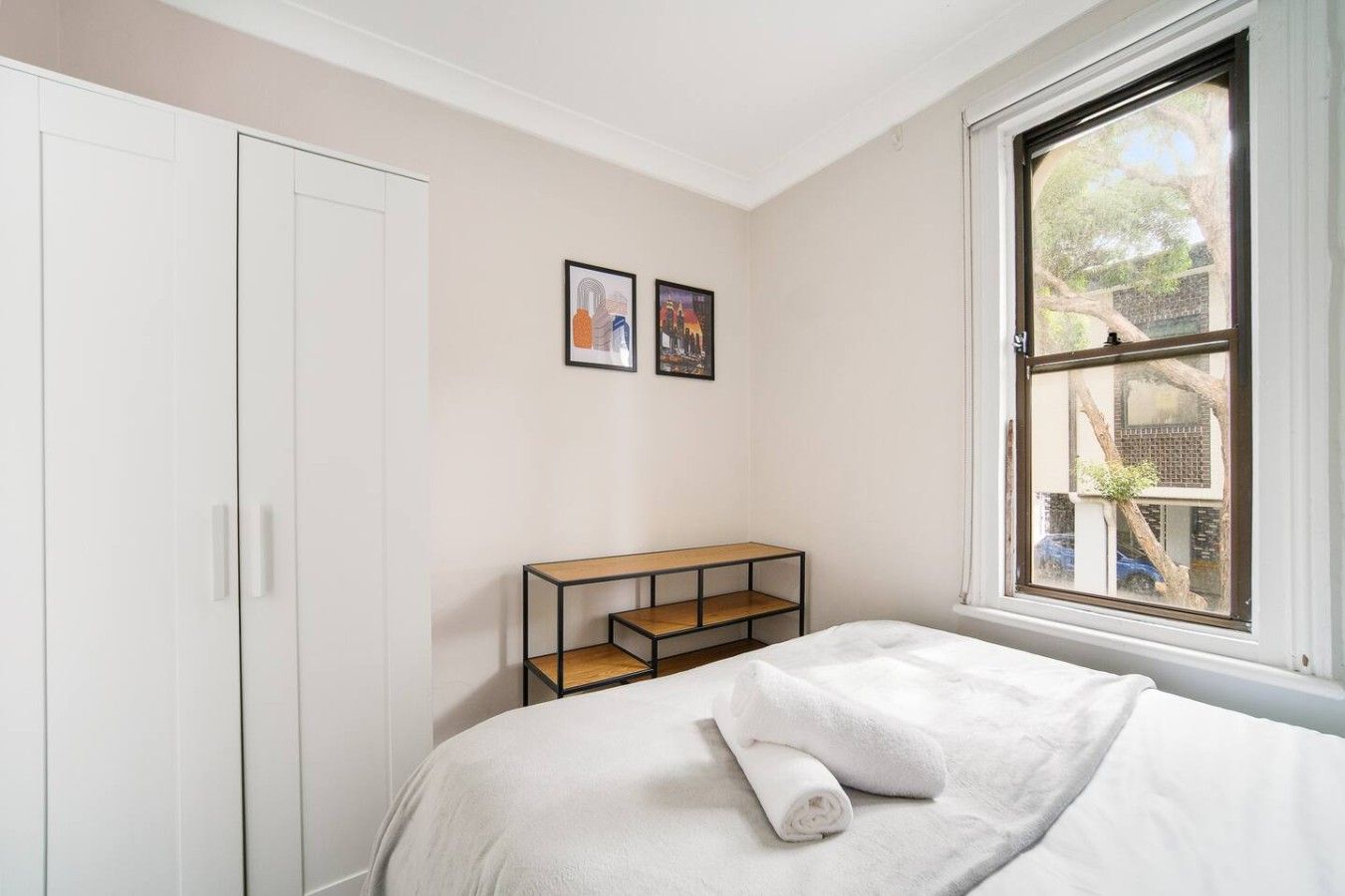A bedroom with a bed, a wardrobe, a window with a view, and a small table with a shelf.