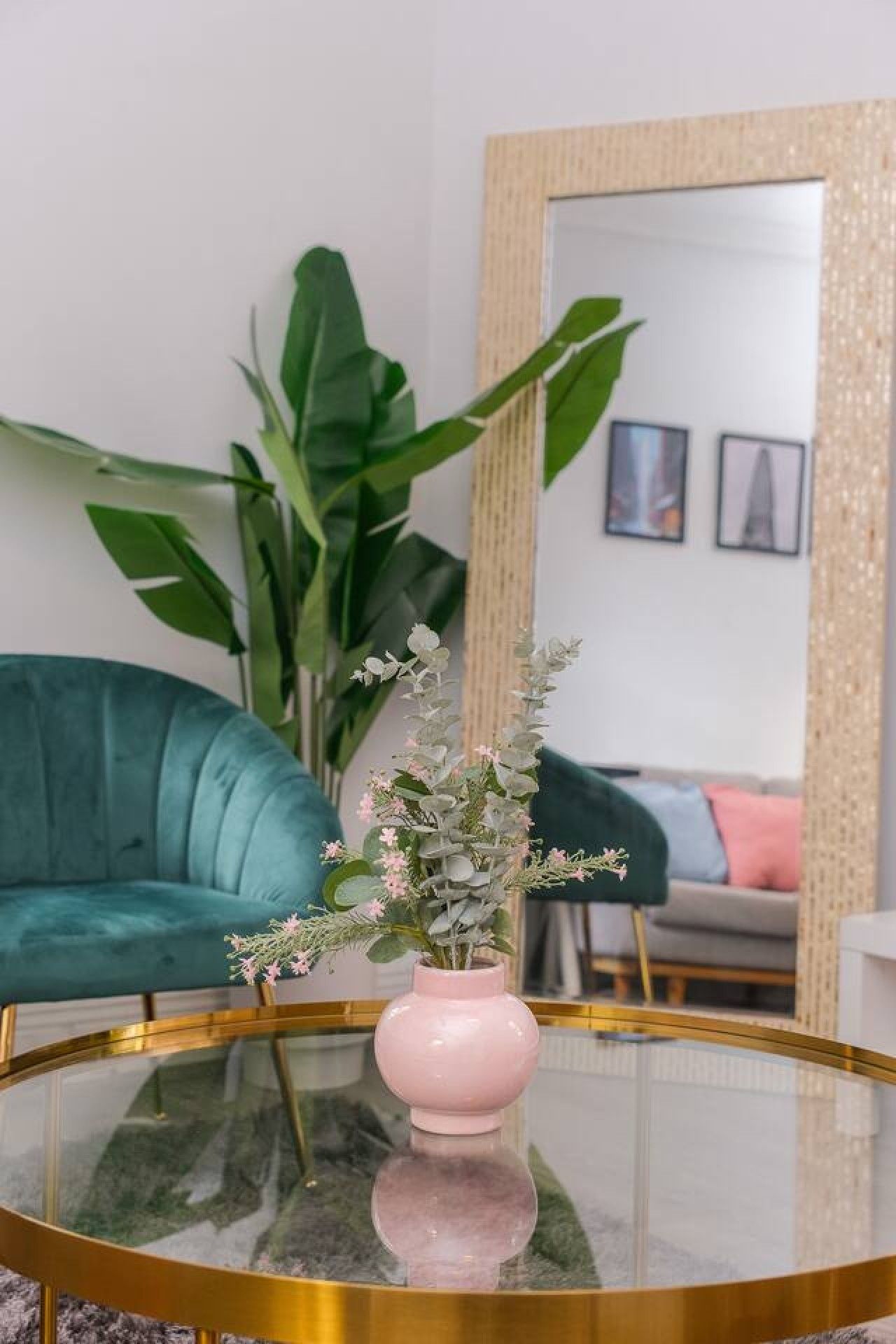 A round glass table with a pink vase containing flowers, a teal sofa, and a large mirror with a decorative frame.