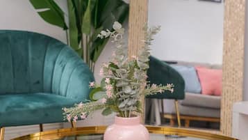 A round glass table with a pink vase containing flowers, a teal sofa, and a large mirror with a decorative frame.