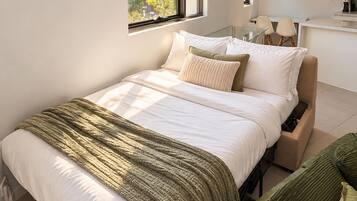 Une chambre moderne avec un lit, des tables de chevet, un canapé vert et une fenêtre donnant sur des immeubles.