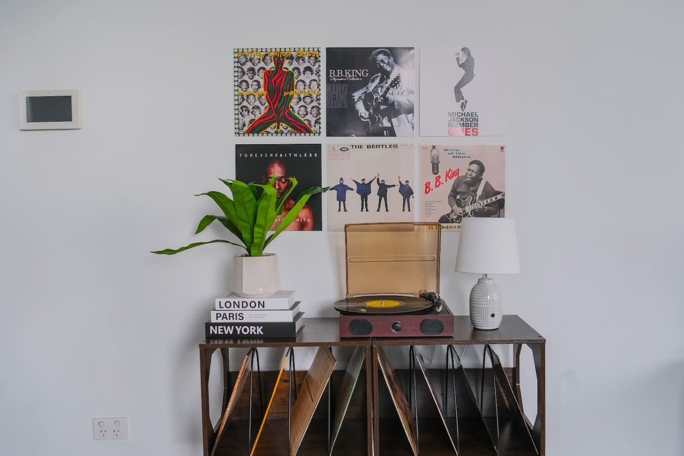 Paredes decoradas com diversos cartazes, um vaso de planta, uma pilha de livros, um toca-discos antigo e um abajur.