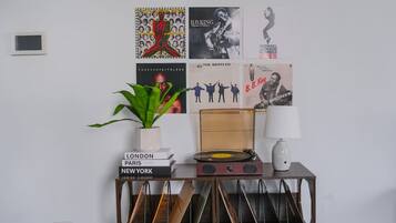 Paredes decoradas com diversos cartazes, um vaso de planta, uma pilha de livros, um toca-discos antigo e um abajur.