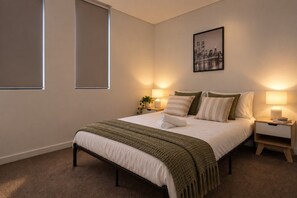A bedroom with a bed, bedside tables, lamps, a cityscape painting, and a window.