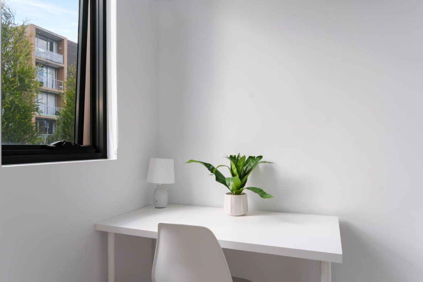 Un escritorio blanco con una silla, una planta en maceta y una lámpara en una esquina junto a una ventana.
