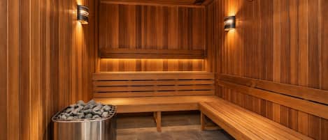 A sauna with wooden benches and walls, a metal container with stones, and wall-mounted lighting.