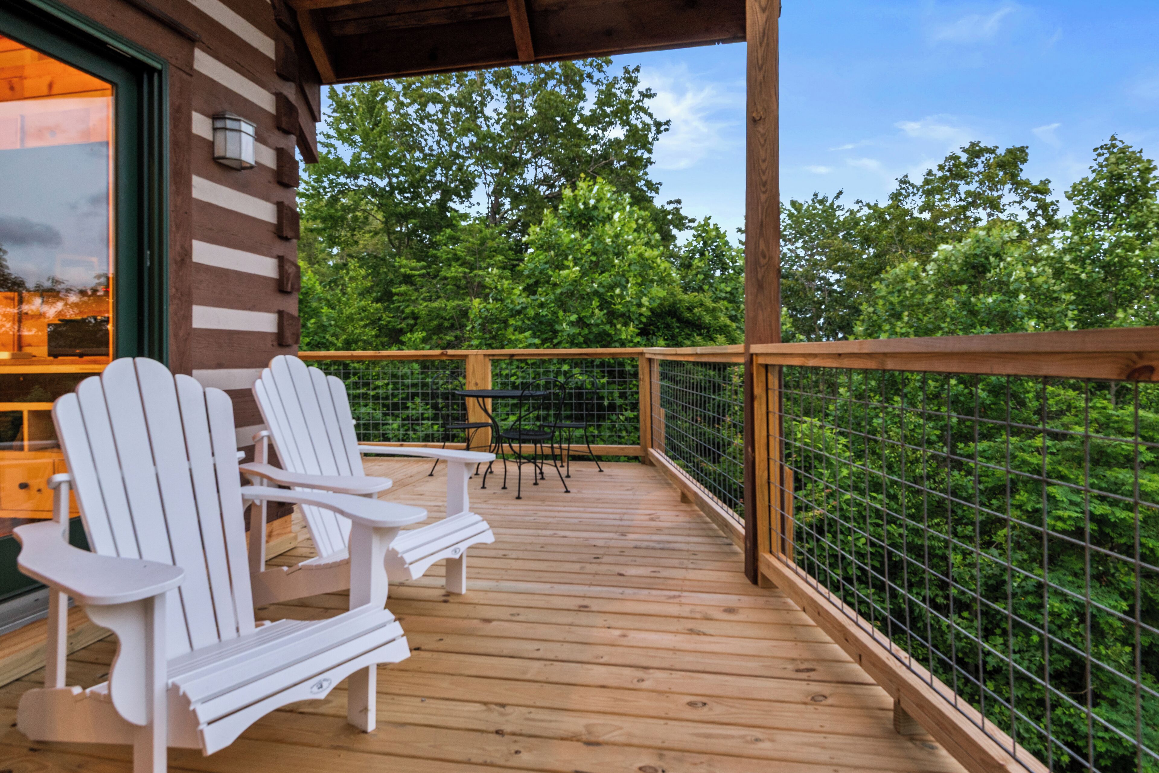 A wooden deck with Adirondack chairs, a table, and a view of a forest.