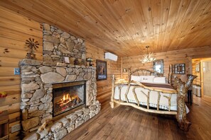A log cabin interior with a stone fireplace, wooden walls, a bed with a unique headboard, and a chandelier. - Lakefront luxury awaits at Beech Mountain Ski and Lake Chalet! (Beech Mountain)