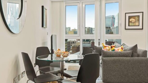 Un salon moderne avec une table à manger en verre, des chaises noires, un canapé gris et une vue sur la ville à travers de grandes fenêtres.