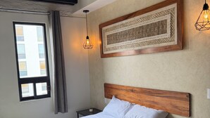 Een netjes opgemaakt bed met een houten hoofdeinde, een nachtkastje met een lamp, een kunstwerk in een lijst aan de muur en een raam met gordijnen.