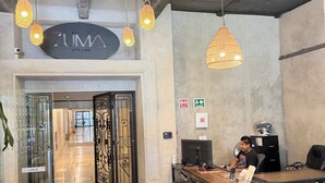 Een ruim interieur met hangende geweven lampen, een receptiebalie en een bord waarop 'ZUMA COLONY' staat.