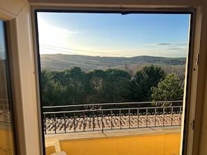 Vista da janela com corrimão de metal, parede amarela e paisagem de árvores e colinas.