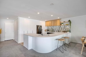 A modern kitchen with a central island, bar stools, a refrigerator, and a wooden cabinet. - Spacious Oceanside Oasis (Mount Maunganui)