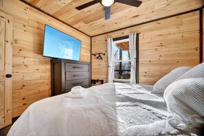 Ein Zimmer in einer Holzhütte mit einem Bett, einem Schminktisch, einem Fernseher und einem Fenster mit Vorhängen.