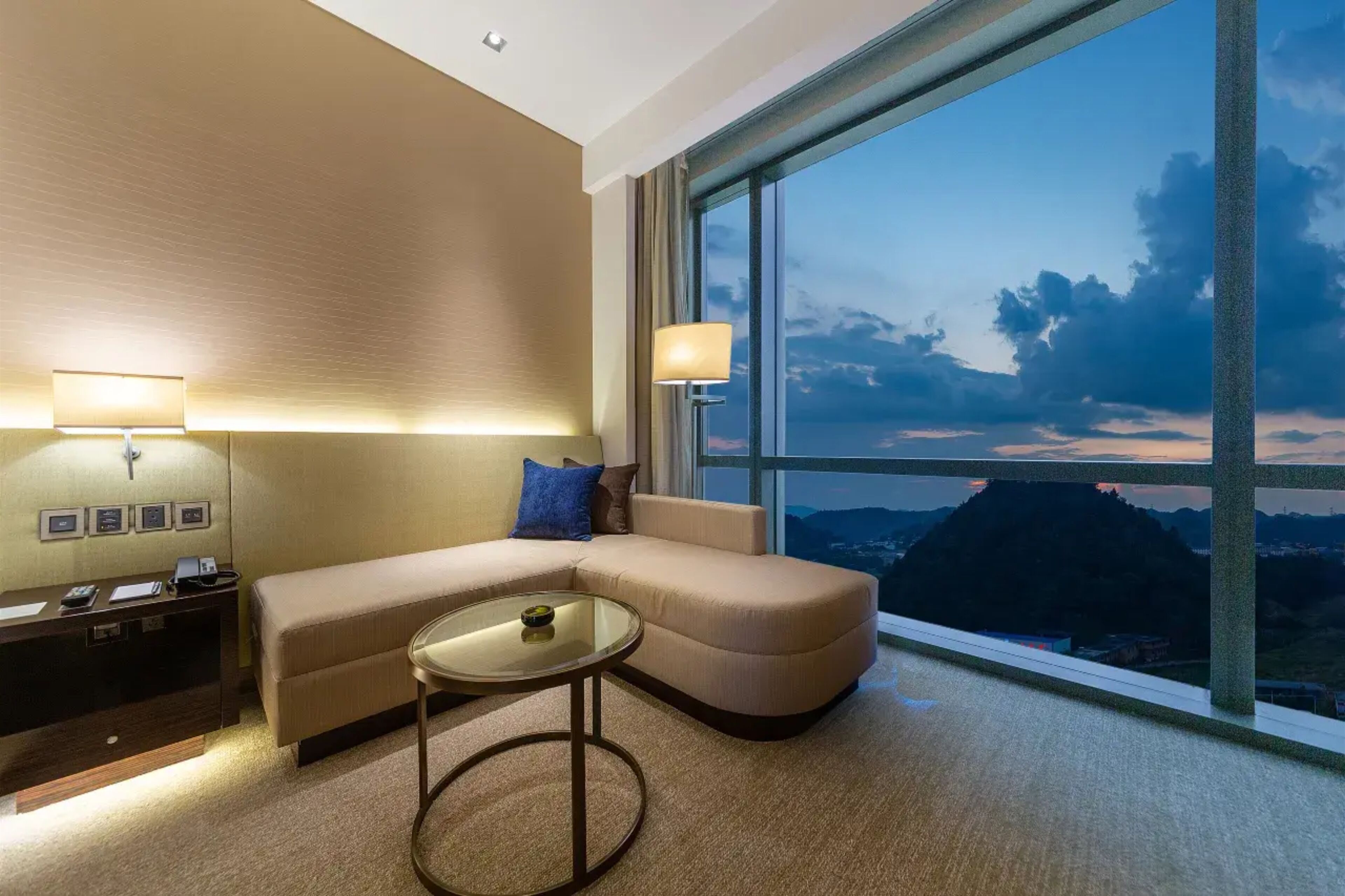 Una habitación de hotel moderna con un ventanal grande, un sofá, una mesita y un escritorio con lámpara.