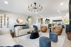 Ein modernes Wohnzimmer mit einer Couch, einem Couchtisch und Sesseln, ein Essbereich mit Tisch und Stühlen sowie eine Küche im Hintergrund.