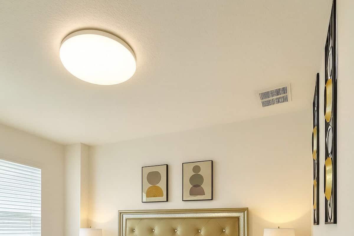 A hotel room with a bed, two bedside tables, a headboard, a ceiling light, a window with blinds, and wall-mounted artwork.