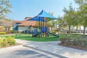 A playground with a slide and climbing structures, a covered seating area, and a building with a red-tiled roof in the background.