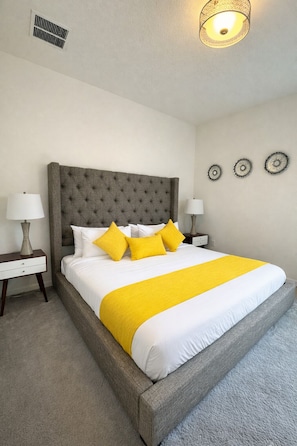 A neatly made bed with a grey headboard, white bedding, and two yellow pillows. There are two bedside tables with lamps, a wall vent, and two wall decorations. - Value Edge (Kissimmee)
