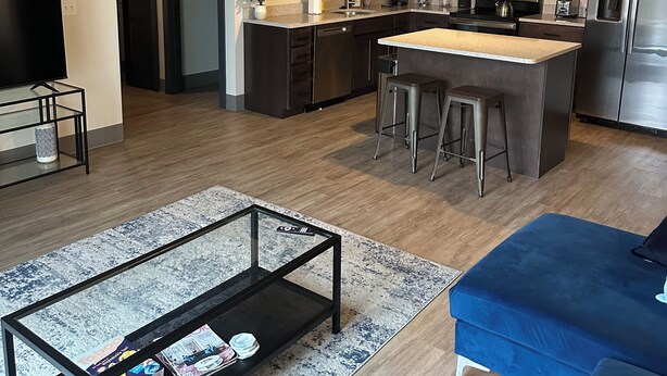 A modern kitchen with stainless steel appliances, a central island with stools, and a dining area with a blue sofa. - Luxury King Suite Downtown Kalamazoo (Kalamazoo)