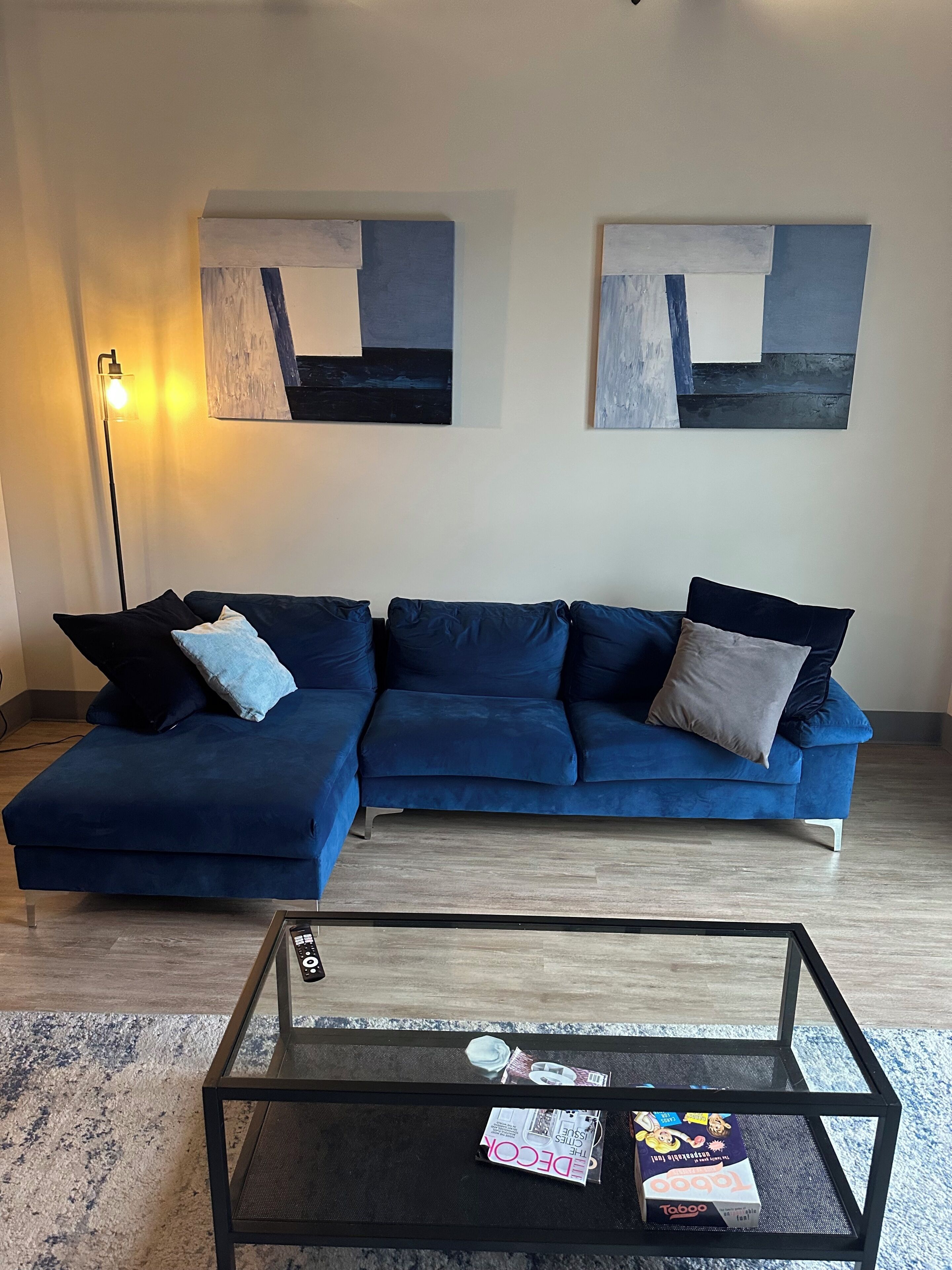 A living room with a blue sectional sofa, a glass coffee table, and abstract wall art.