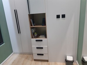 Una habitación con un armario, una pequeña estantería con libros y una planta, además de un cubo de basura.