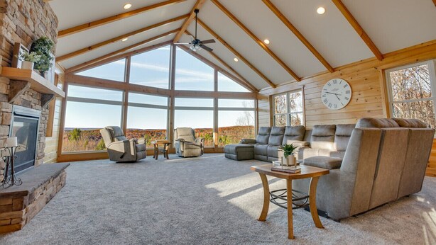 Ein geräumiges Wohnzimmer mit Kamin aus Stein, großen Fenstern, einem Deckenventilator und einer Wanduhr.