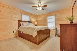 A bedroom with a wooden bed, a ceiling fan, two windows with blinds, and a small plant on a wooden cabinet.