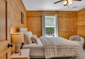 Ein Schlafzimmer mit Holzwandverkleidung, einem Bett, einer Nachttischlampe, einem Fenster mit Jalousien und einem Deckenventilator.