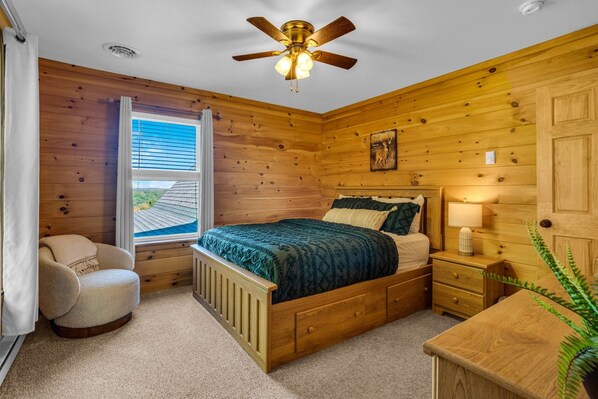 A wooden cabin bedroom with a bed, a chair, a nightstand, a lamp, and a window with blinds.