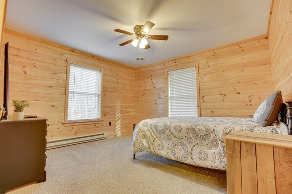A bedroom with wooden walls, a bed with patterned bedding, a ceiling fan, and two windows with blinds.