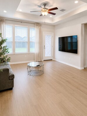 A spacious living room with a ceiling fan, a large TV, and a dining table. - Modern Sunfield Home w/ Pool & Gym (Buda)