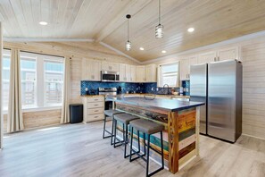 A modern kitchen with wooden cabinets, a large island with stools, stainless steel appliances, and a view of the city. - CL6 Shared Pool, Charcoal Grill, Old Town Port A (Port Aransas)