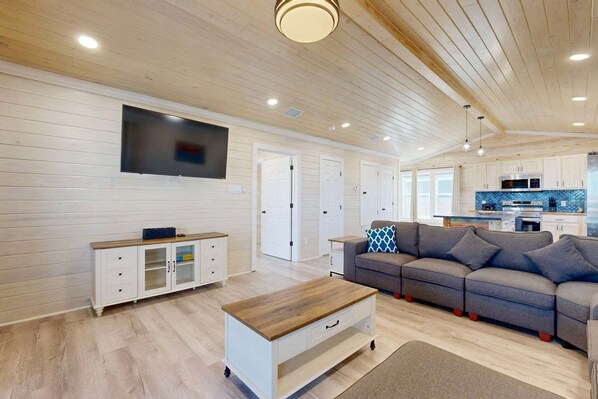 A spacious living room with a wooden ceiling, a flat-screen TV mounted on the wall, a grey sectional sofa, a white cabinet with drawers, and a wooden coffee table. - CL6 Shared Pool, Charcoal Grill, Old Town Port A (Port Aransas)
