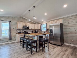 A modern kitchen with wooden cabinets, a stainless steel refrigerator, a central island with stools, and pendant lighting. - CL8 Shared Pool, Charcoal Grill, Old Town Port A (Port Aransas)