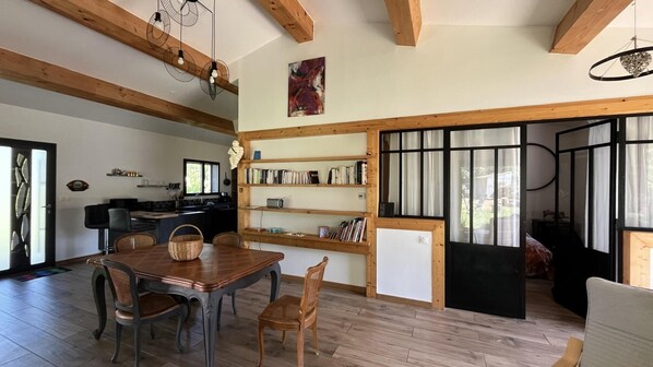 A dining area with a wooden table and chairs, a kitchen with a window, and a bookshelf.