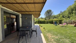 An outdoor patio with a table and chairs, a covered area, and a well-maintained lawn with trees and shrubs.