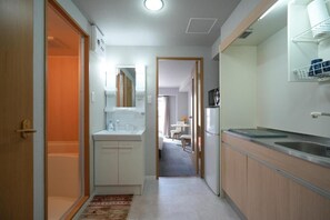 A narrow hallway leading to a bathroom with a toilet and sink, a small kitchen area with a refrigerator and microwave, and a dining area visible through an open door. - Dc Sakura Hotel - Namba Branch (Osaka)