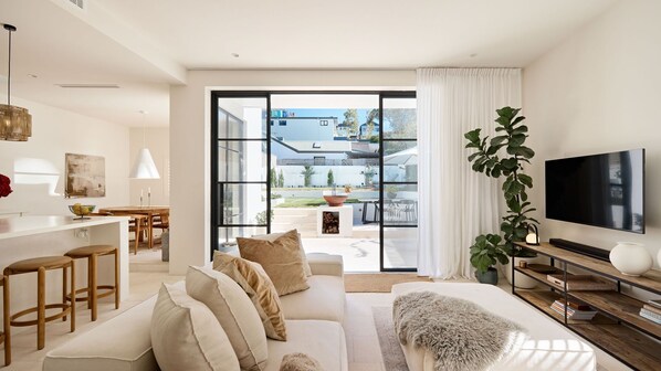 A modern living room with a sofa, TV, and a large window overlooking a garden. - Casa Bianca, Bondi IH (Bondi)