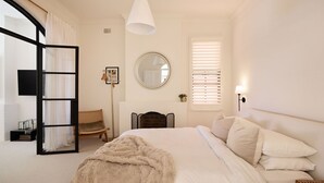 A bedroom with a bed, a chair, a fireplace, a mirror, and a window with blinds. - Casa Bianca, Bondi IH (Bondi)