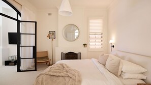 A bedroom with a bed, a chair, a fireplace, a mirror, and a window with blinds. - Casa Bianca, Bondi IH (Bondi)