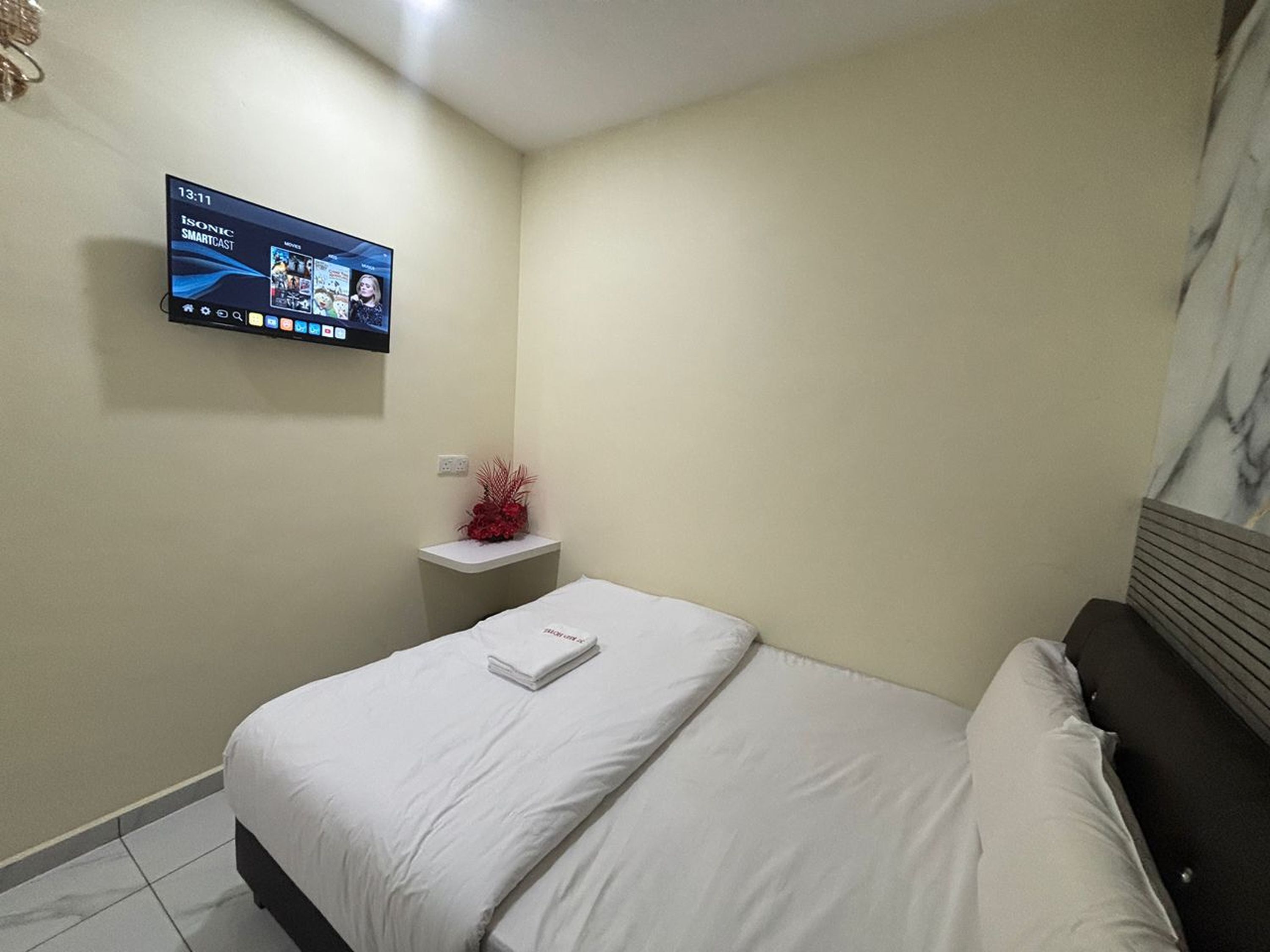 Habitación de hotel con cama, televisor montado en la pared, una mesita de noche pequeña con un adorno rojo y pared con diseño de mármol.
