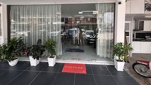 Entrada de un hotel con felpudo rojo, plantas en macetas y vista de coches aparcados a través de las puertas de cristal.