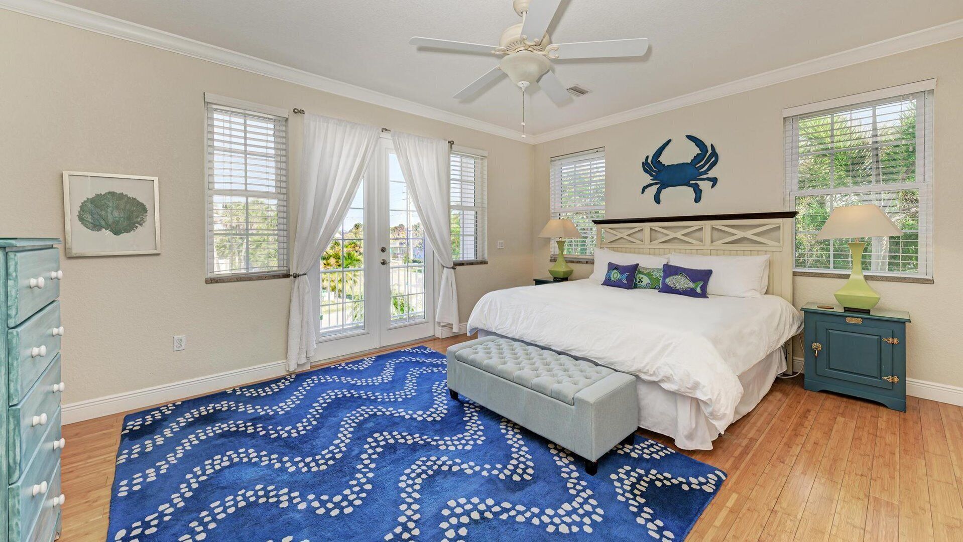 A bedroom with a bed, a ceiling fan, a window with curtains, a wall-mounted crab decoration, a nightstand with a lamp, and a blue patterned rug.