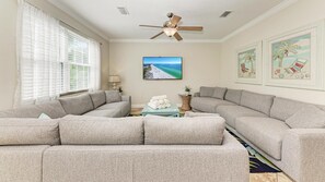 A living room with a sectional sofa, a ceiling fan, a TV, and beach-themed artwork. - Casa Morado | Luxury Holmes Beach Retreat with Heated Pool and Hot tub, Steps to Beach  I .PET FRIENDLY. ANNA MARIA ISLAND! (Holmes Beach)