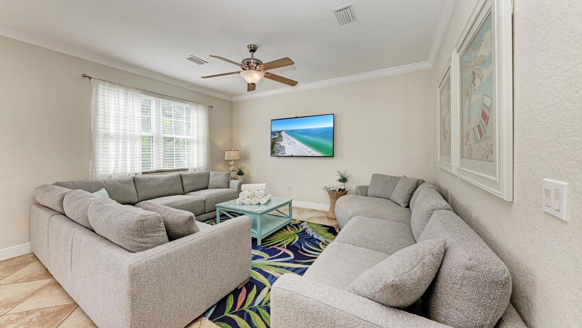 A living room with a sectional sofa, a ceiling fan, a flat-screen TV, and a beach-themed painting.